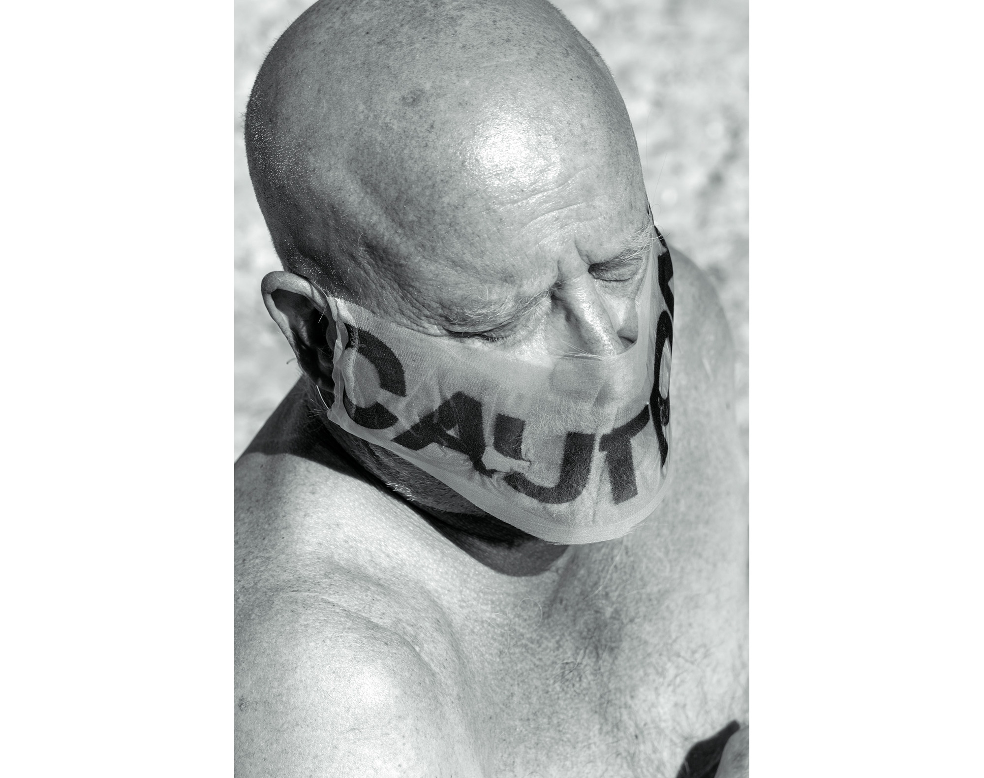 Black and white photograph of a bare-chested bald man with shaggy eyebrows wearing a transparent gauze face mask with the word ‘caution’ printed across the front, covering his nose, lips, and mouth. Details of lips and short gray beard can be seen through the fabric and lettering on the mask.